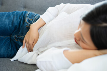 Unhappy Asian woman sitting on the sofa and holding on stomach suffering. Abdominal pain that comes...