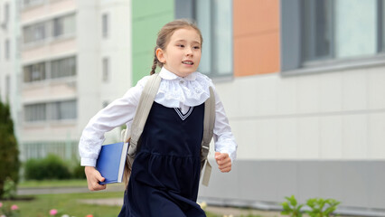 Junior schoolgirl holding blue book runs to elementary school. Long-haired girl with backpack late...