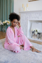 Young african woman in pink pajamas sitting on carpet in bedroom.