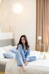 Young pensive brunette woman in blue pajamas sitting on bed.