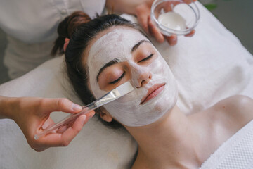 Top view of crop anonymous female applying facial mask on face while sitting on bed with closed eyes