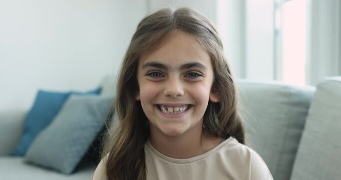Close up head shot cute little girl sit on sofa smile look pose for camera. Video call, virtual meeting event participant or blogger, dental clinic services for children, alpha generation kid portrait