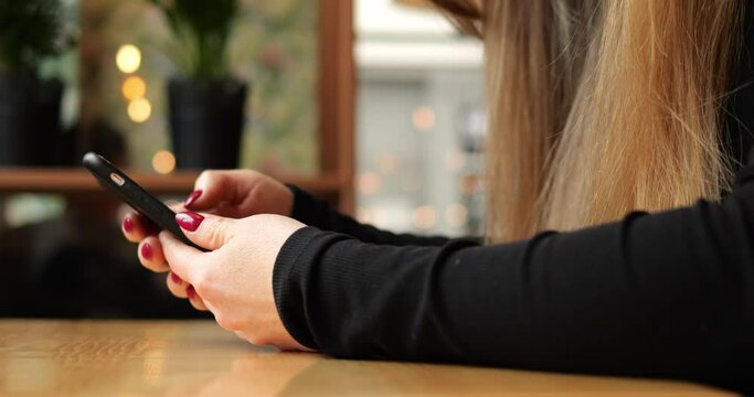 Close up woman arm hold smartphone lie on wooden table inside public building. Hands of anonymous female with long brown hair and red nail polish in black long sleeve with mobile phone.