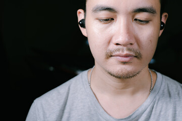 Obraz premium Close-up shot of a partial face of a young Asian man wearing a grey t-shirt listening to music on his wireless earphones. Shot from front of him. Isolated black background.