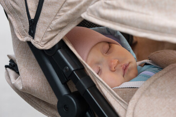 Toddler girl falls asleep in baby cradle during walking with mother in city park. Child in pink hat sleeps seeing picturesque dreams