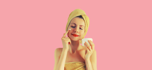 Obraz na płótnie Canvas Portrait of happy smiling young caucasian woman applying face cream and drying wet hair in wrapped bath towel on pink background