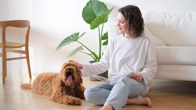 Dog chilling with owner. Young woman in blue jeans reading book while sitting on floor at home petting labradoodle poodle dog. Weekend time for hobby, slow living concept, cozy lifestyle in city flat.