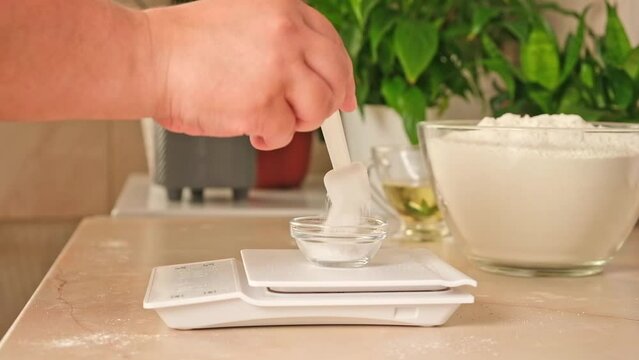 A woman measures salt into a glass bowl standing on an electronic scale. Preparation of yeast dough for pizza.