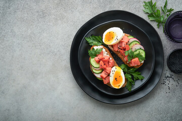 Healthy sandwich with bread or toast, smoked salmon, soft egg, cream cheese, cucumber, radish, black sesame and parsley on black plate. Delicious protein fish sandwich for breakfast. Smorrebrod.