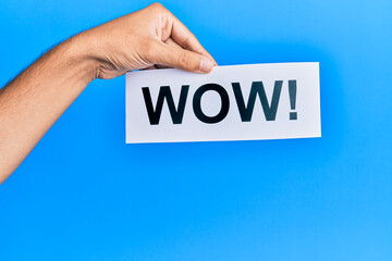 Hand of caucasian man holding paper with wow! message over isolated blue background