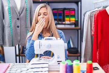 Blonde woman dressmaker designer using sew machine bored yawning tired covering mouth with hand....