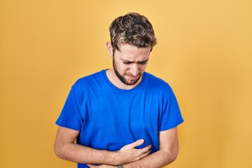 Hispanic man with beard standing over yellow background with hand on stomach because indigestion,...