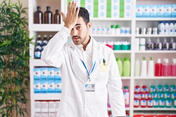 Handsome hispanic man working at pharmacy drugstore surprised with hand on head for mistake, remember error. forgot, bad memory concept.