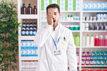 Handsome hispanic man working at pharmacy drugstore yawning tired covering half face, eye and mouth...