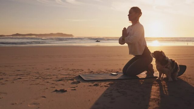 Woman doing workout with dog on ocean beach at sunset. Female doing Yoga and stretching on mat on sand sea shore at sunrise. Strong powerful woman exercises in the morning or evening outdoors. 