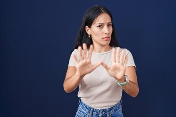 Young hispanic woman standing over blue background moving away hands palms showing refusal and denial with afraid and disgusting expression. stop and forbidden.