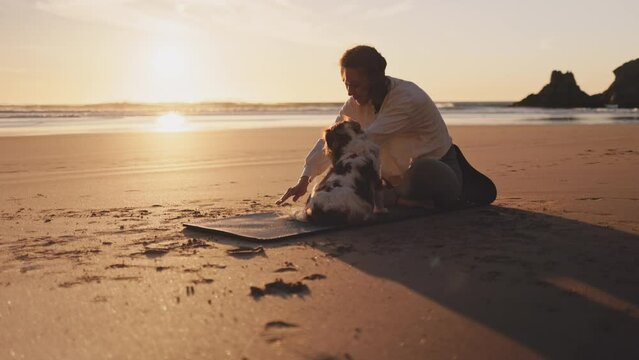 Woman doing workout with dog on ocean beach at sunset. Female doing Yoga and stretching on mat on sand sea shore at sunrise. Strong powerful woman exercises in the morning or evening outdoors. 