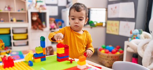 Adorable hispanic boy playing with construction blocks standing at kindergarten