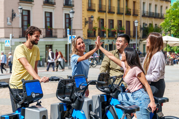 A group of five diverse, happy friends high-fiving while renting electric bikes on a sunny day in the city.