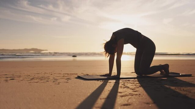 Woman doing workout with dog on ocean beach at sunset. Female doing Yoga and stretching on mat on sand sea shore at sunrise. Strong powerful woman exercises in the morning or evening outdoors. 