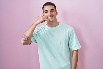 Handsome hispanic man standing over pink background smiling doing phone gesture with hand and fingers like talking on the telephone. communicating concepts.