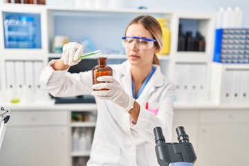 Young hispanic woman scientist pouring liquid on bottle at laboratory