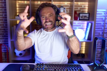 Middle age man with beard playing video games wearing headphones shouting frustrated with rage, hands trying to strangle, yelling mad