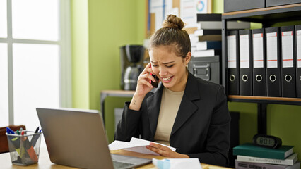 Young beautiful hispanic woman business worker reading letter with surprise expression talking on smartphone at office