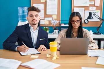Young hispanic man and woman working at the office thinking attitude and sober expression looking self confident