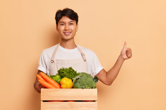 Young farm guy poses with organic veg box on peachy background in studio, smiles and shows thumb up, organic food concept, copy space