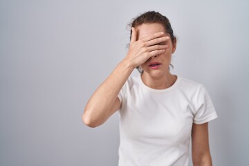 Beautiful brunette woman standing over isolated background covering eyes with hand, looking serious...