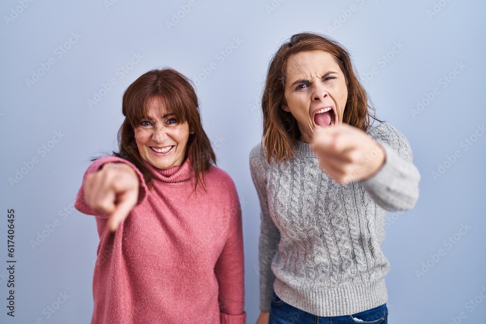 Poster mother and daughter standing over blue background pointing displeased and frustrated to the camera, 