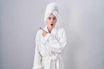 Blonde caucasian woman wearing bathrobe surprised pointing with finger to the side, open mouth...