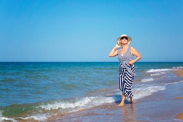 Smiling elderly woman in a dress and hat walks along the sandy beach near the sea on a sunny day. Active recreation, travel and freedom.