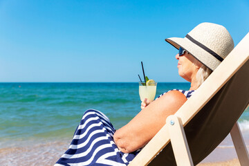 A smiling elderly woman in a dress and hat sits in a sun lounger on the seashore with a drink in her hands. Active lifestyle, travel and freedom.