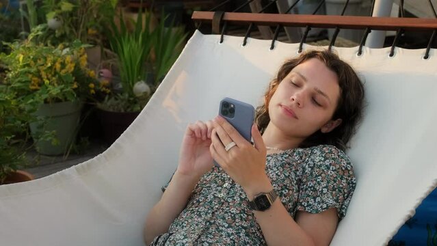 Portrait young curly woman lies in hammock use mobile phone online surrounded by garden flowers. Smile female in floral print sundress relaxes on her home terrace after working day enjoys sunset light