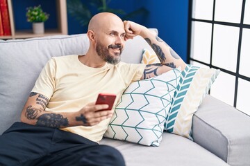 Fototapeta premium Young bald man using smartphone sitting on sofa at home