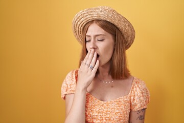 Young redhead woman standing over yellow background wearing summer hat bored yawning tired covering mouth with hand. restless and sleepiness.