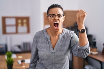 African american woman working at the office wearing glasses angry and mad raising fist frustrated and furious while shouting with anger. rage and aggressive concept.