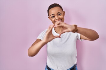 Beautiful african american woman standing over pink background smiling in love doing heart symbol shape with hands. romantic concept.