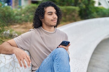 Obraz premium Young latin man using smartphone sitting on bench at park