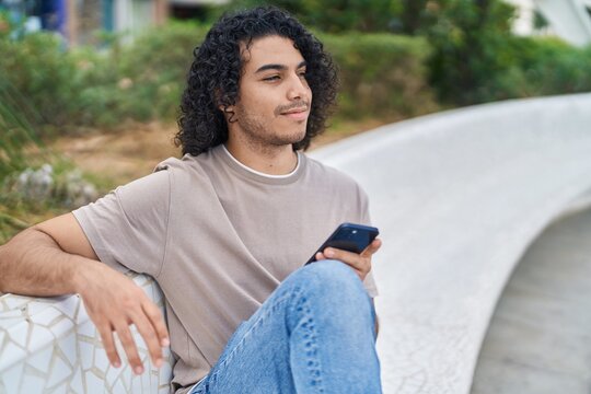 Young latin man using smartphone sitting on bench at park