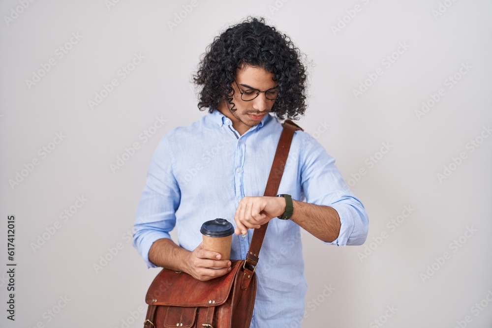 Wall mural Hispanic man with curly hair drinking a cup of take away coffee checking the time on wrist watch, relaxed and confident