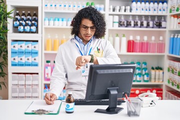 Hispanic man with curly hair working at pharmacy drugstore looking at the watch time worried,...