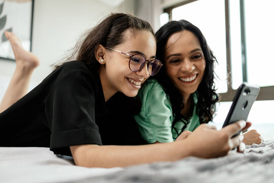 Mother And Teenage Daughter Using Smartphone Together Lying In Bed At Home. Latina Mother And Daughter Looking At Phone Watching Social Media Videos While Relaxing In Bedroom. Family And Technology