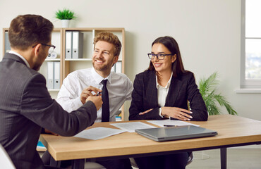 Happy, young couple sitting at office desk with credit manager, realtor or financial advisor. Estate agent, loan broker or business adviser offers clients a contract and gives pen to sign it