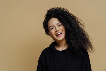 Positive African American woman smiles, tilts head happily, wearing black sweatshirt, isolated on...