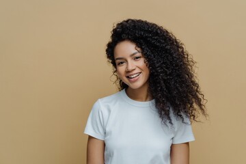 Beautiful woman with frizzy hair, gentle smile, healthy skin, casual white t-shirt, conversing, beige background. People, emotions. - obrazy, fototapety, plakaty