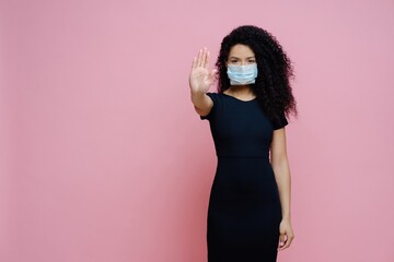 Ethnic woman with curly hair, making stop gesture, says no to COVID-19. Wearing mask, black dress,...