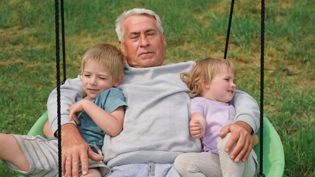 Grandfather swinging children in summer park. Grand dad and grandchildren sitting on swing outdoors. Senior 60s Grandpa pushing small grandkids on a rope seesaw. Old man and little kids at playground